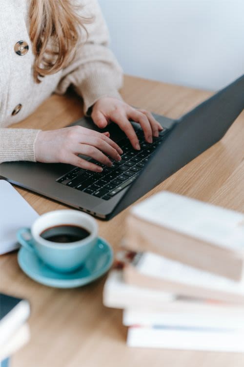Woman working at a laptop with a coffee and pile of books next to her