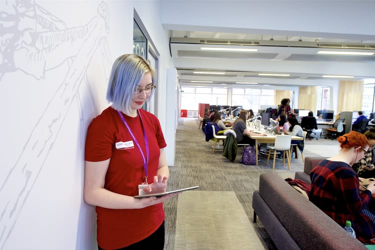 A member of the Student Team stands against a wall in AGLC looking at a tablet device
