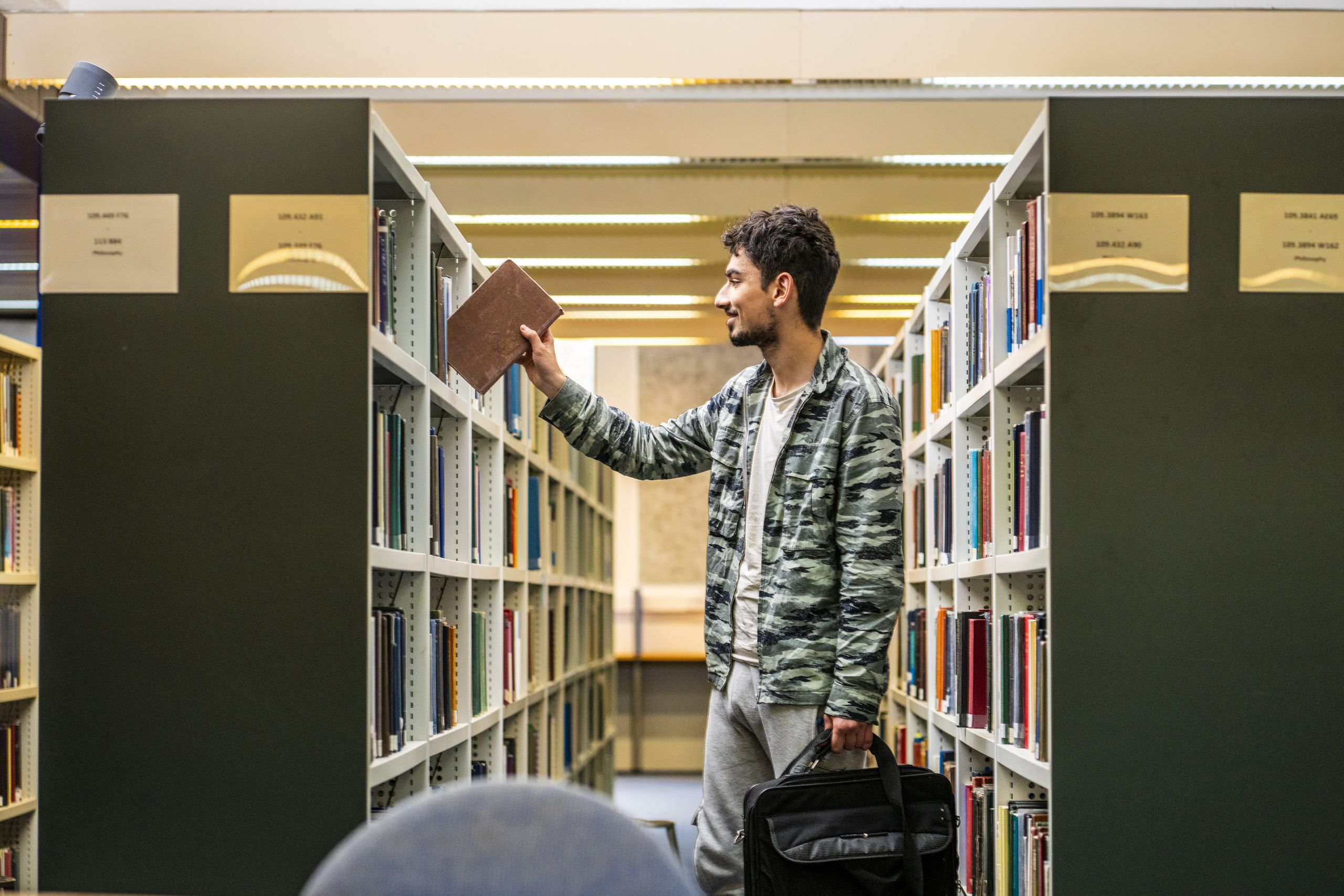 Student taking a book off a shelf while holding a laptop bag