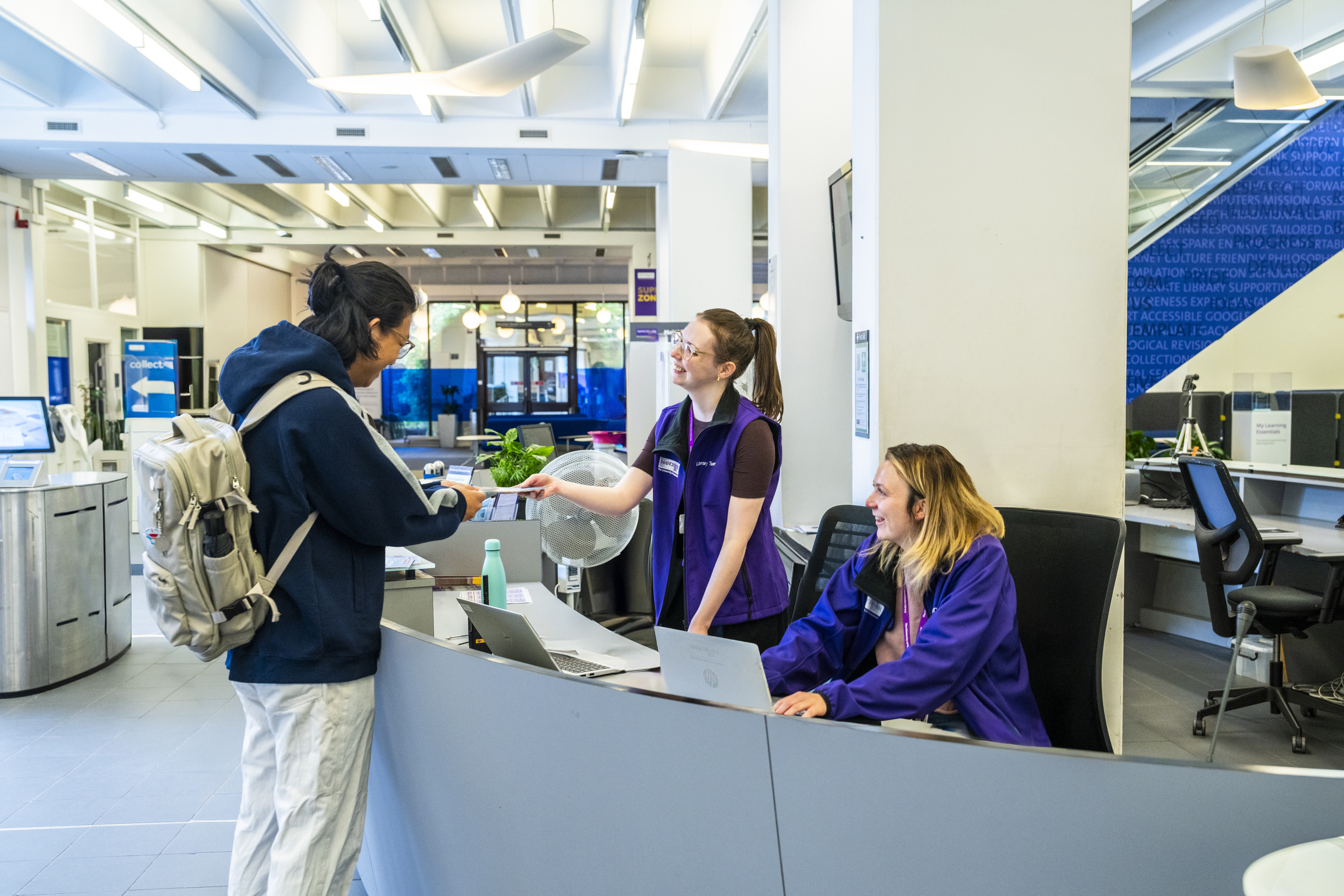 Student smiling with Library staff who are behind the service desk in Main Library