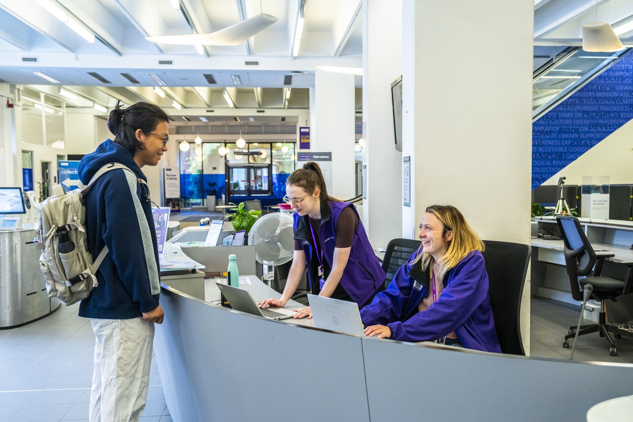 Student smiling with Library staff who are behind the service desk in Main Library