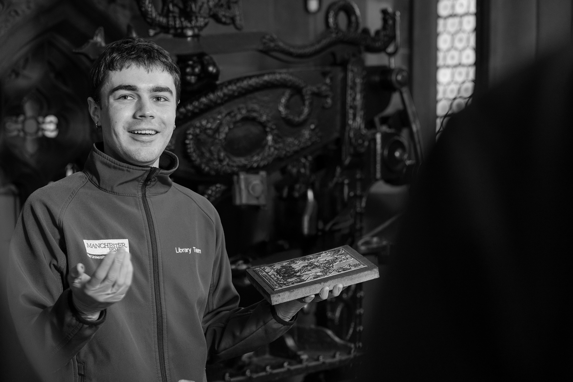 Member of Rylands staff holding an item from collections in front of a printing press