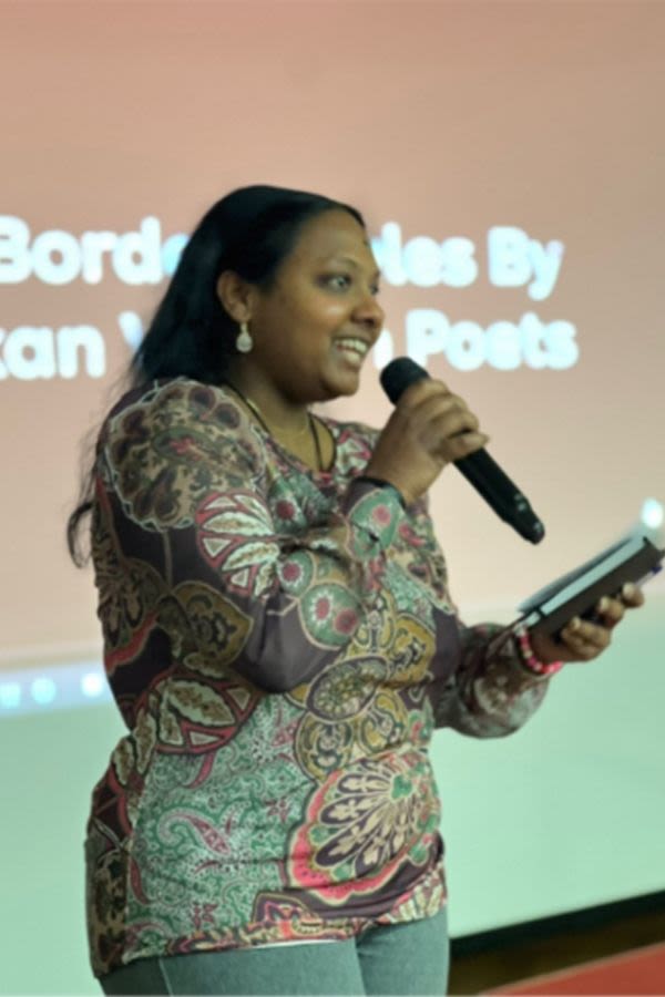 A woman reading from a book holding a microphone on stage in front of Powerpoint slides