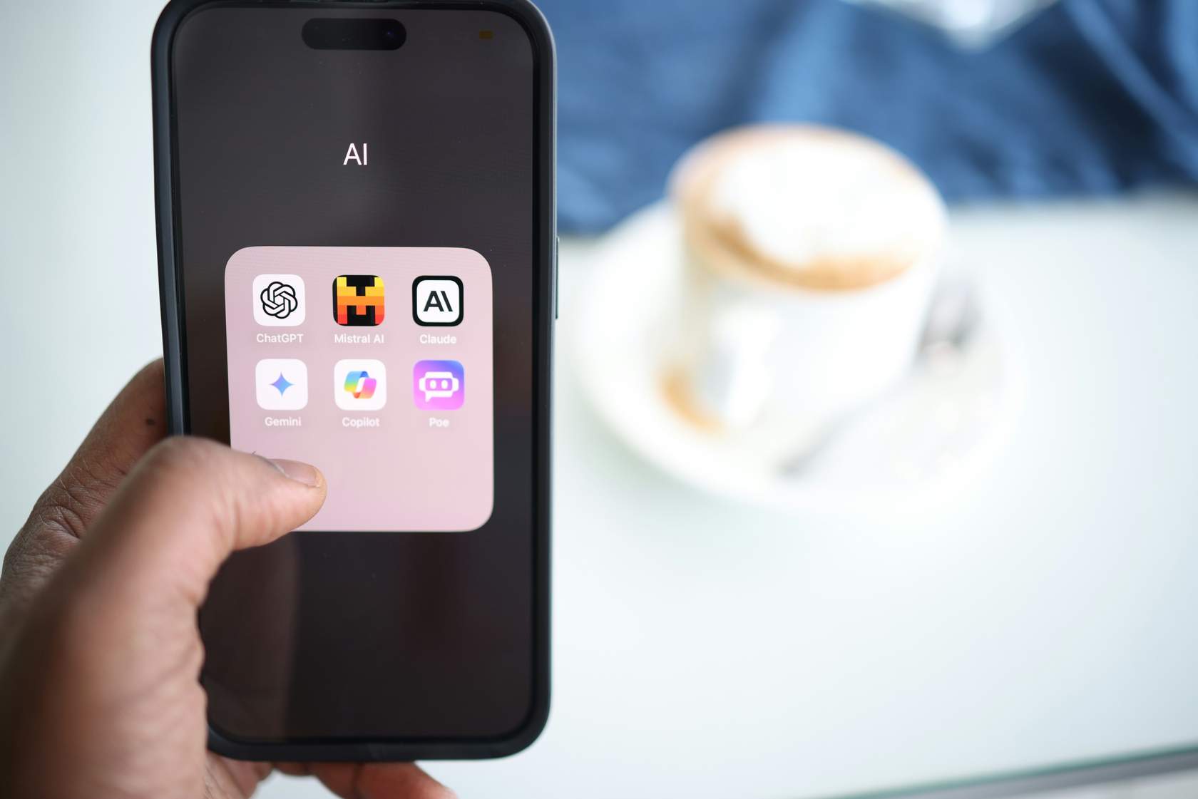 A phone held up in front of a cup of coffee displaying logos for AI apps