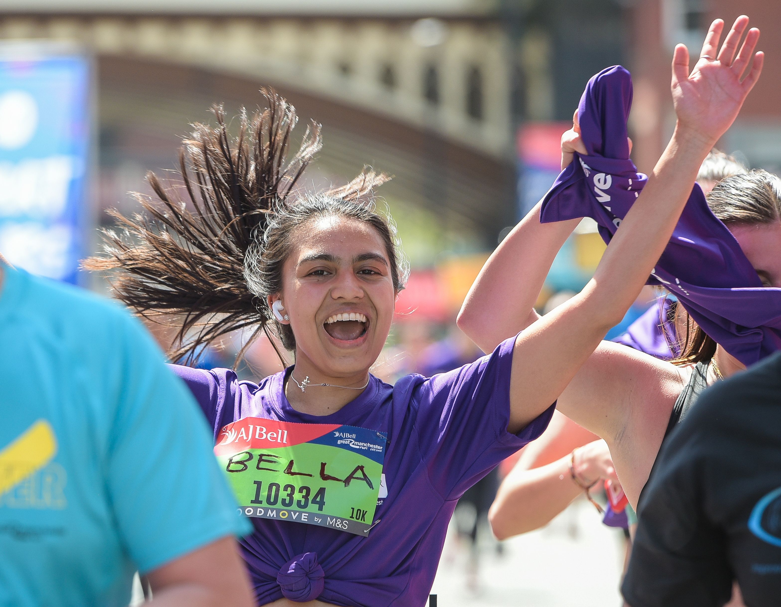 A woman wearing a purple t-shirt crosses the finish line of a race. She's smiling broadly and her arms are in the air in a gesture of triumph. 